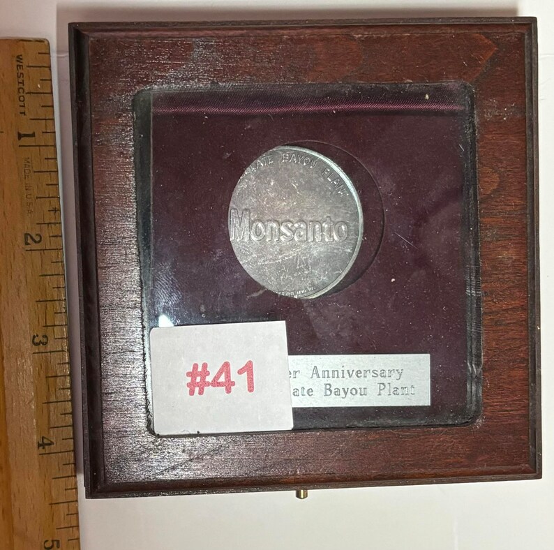 May include: A silver coin with the text "Monsanto" and "Gate Bayou Plant" in a wooden display case with a red velvet lining. The case has a label with the number "#41" and the text "41st Anniversary".