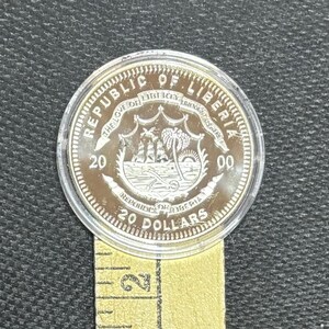 May include: A silver coin from the Republic of Liberia, featuring a detailed emblem with a ship, palm trees, and the text "20 Dollars." The coin is encased in a clear plastic holder and rests on a yellow measuring tape.