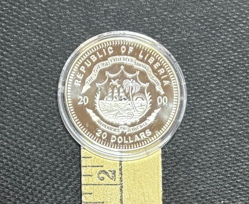 May include: A silver coin from the Republic of Liberia, featuring a detailed emblem with a ship, palm trees, and the text "20 Dollars." The coin is encased in a clear plastic holder and rests on a yellow measuring tape.