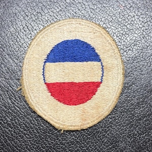 May include: A circular embroidered patch featuring a tan background with a central round emblem. The emblem consists of three horizontal stripes in blue, tan, and red, enclosed by a thin blue border, all stitched with a textured thread finish.