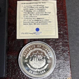 May include: A silver coin from the Republic of Liberia, featuring a ship and palm trees, with the text "20 DOLLARS". The coin is displayed in a protective case with a guarantee document from the American Mint.