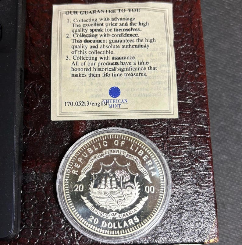 May include: A silver coin from the Republic of Liberia, featuring a ship and palm trees, with the text "20 DOLLARS". The coin is displayed in a protective case with a guarantee document from the American Mint.