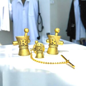 May include: Three gold-toned cufflinks in the shape of a mortar and pestle with the symbol "Rx" on the side. The cufflinks are on a white surface.