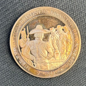May include: A bronze-colored commemorative coin with the text "Mier Expedition Prisoners Drawing Black Beans" encircling a detailed relief. The central image depicts figures in period clothing, with the year 1843 at the bottom. The coin has a polished, reflective surface.