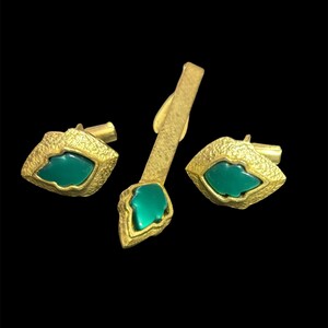 May include: A set consisting of two cufflinks and a tie bar, each featuring a textured gold-tone metal finish and a central, smooth, leaf-shaped green stone inlay. The cufflinks have a geometric shape, while the tie bar is elongated.