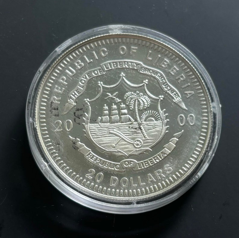 May include: Silver coin from the Republic of Liberia, featuring the country's coat of arms with a palm tree, a ship, and a sunrise. The coin is dated 2000 and has a face value of 20 dollars.