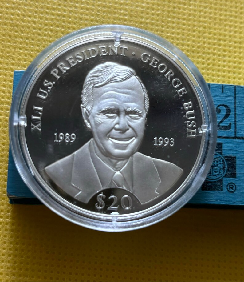 May include: A silver commemorative coin featuring a portrait of George H.W. Bush, the 41st U.S. President. The coin reads "XLI U.S. PRESIDENT GEORGE BUSH" and includes the years "1989" and "1993". The coin also features the denomination "$20".