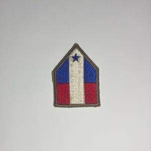 May include: A pentagonal embroidered patch featuring a vertical white stripe centered between blue and red sections. A small blue star is stitched at the top of the white stripe. The entire patch is outlined with a thick, dark brown border.