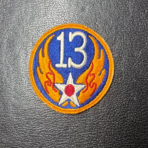 May include: A circular embroidered patch featuring a blue background with a yellow border. The design includes the white number 13 above a white star with a red center, flanked by two stylized yellow and orange flame-like wing shapes.