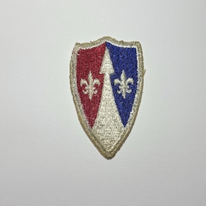 May include: An embroidered shield-shaped patch featuring a central white vertical arrow dividing a red section on the left and a blue section on the right. Each colored section contains a white fleur-de-lis symbol, all bordered by a tan edge.