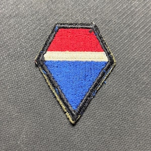 May include: A diamond-shaped embroidered patch featuring a horizontal design with a red top section, a thin white middle stripe, and a blue bottom section. The entire patch is outlined with a thick black stitched border on a textured surface.