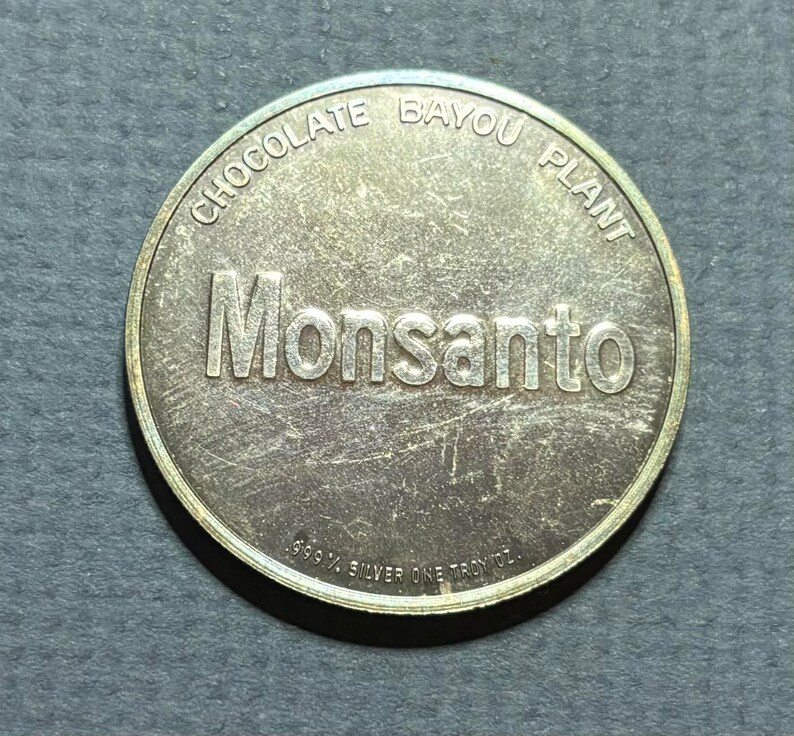 May include: A silver coin with the words "Chocolate Bayou Plant" and "Monsanto" inscribed around the edge. The coin also reads "99.9% Silver One Troy oz."