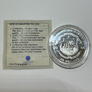 May include: A silver coin with a depiction of a ship sailing in front of a palm tree on a shield. The coin is inscribed with "Republic of Liberia" and "20 Dollars".