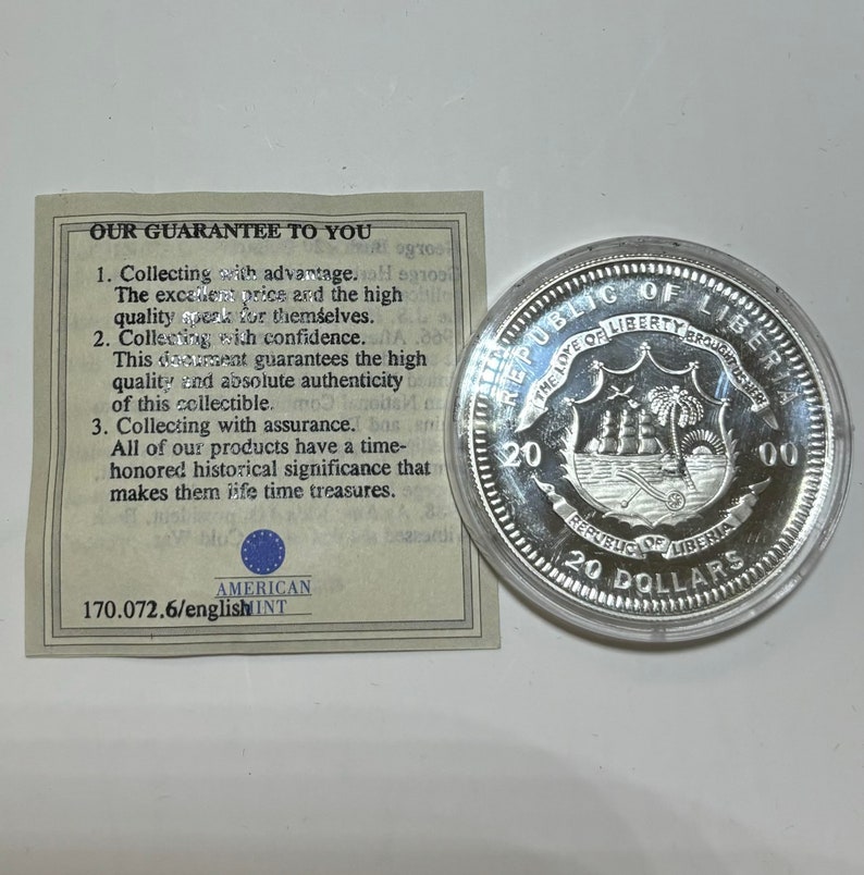 May include: A silver coin with a depiction of a ship sailing in front of a palm tree on a shield. The coin is inscribed with "Republic of Liberia" and "20 Dollars".