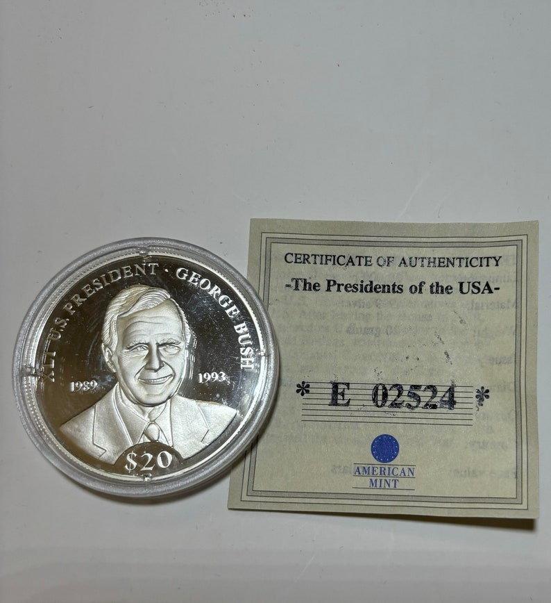 May include: A silver coin with a portrait of George H.W. Bush, the 41st President of the United States. The coin is inscribed with "41st U.S. President George Bush" and the years "1989" and "1993". The coin is also inscribed with "$20". The coin is accompanied by a certificate of authenticity from the American Mint.