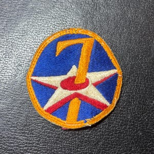 May include: A circular embroidered patch featuring a blue background with a central white star outlined in red. A large, vertical orange number seven is stitched over the center of the star. The entire patch is bordered by a thick orange thread edge.