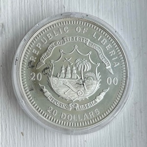 May include: Silver coin from the Republic of Liberia, featuring the country's coat of arms with a ship, palm tree, and cannon. The coin is dated 2000 and reads "20 Dollars".