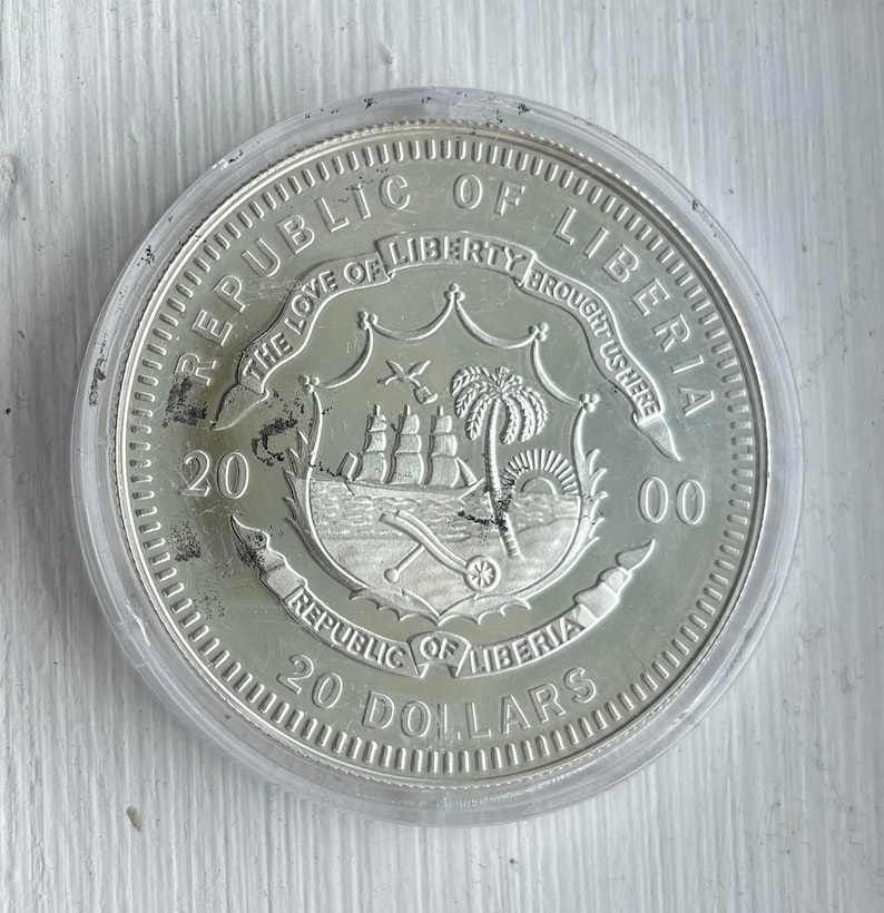 May include: Silver coin from the Republic of Liberia, featuring the country's coat of arms with a ship, palm tree, and cannon. The coin is dated 2000 and reads "20 Dollars".