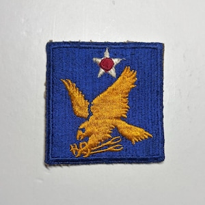 May include: A square embroidered patch featuring a blue background with a golden eagle in the center. The eagle is clutching a pair of golden scissors in its talons. Above the eagle is a white five-pointed star with a red circular center.