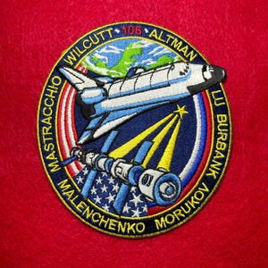 May include: Embroidered patch featuring a space shuttle, a satellite, and the Earth. The patch is circular with a blue, white, and red color scheme, and includes the names of astronauts. The background is red.