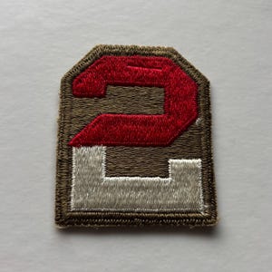 May include: A shield-shaped embroidered patch featuring a brown background with a large number two. The top half of the number is stitched in red thread, while the bottom half is stitched in white thread. The entire patch has a brown stitched border.
