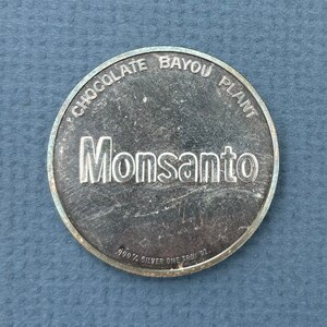 May include: A silver coin with the words "Chocolate Bayou Plant" and "Monsanto" engraved on it. The coin also says "99.9% Silver One Troy Oz."