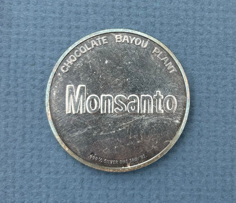 May include: A silver coin with the words "Chocolate Bayou Plant" and "Monsanto" engraved on it. The coin also says "99.9% Silver One Troy Oz."
