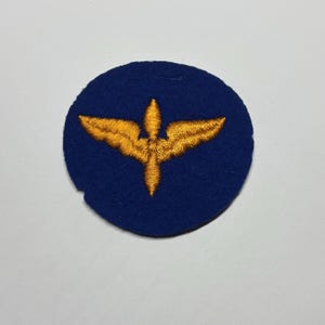 May include: A circular blue embroidered patch featuring a centered golden yellow winged emblem. The design consists of two symmetrical wings extending horizontally from a central vertical point, all rendered in dense, textured thread stitching.