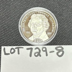 May include: A silver coin depicting Thomas Jefferson, with the inscription "III US PRESIDENT THOMAS JEFFERSON". The coin includes the years 1801 and 1809, and the value $20. Below the coin is a white paper with the text "LOT 729-8".