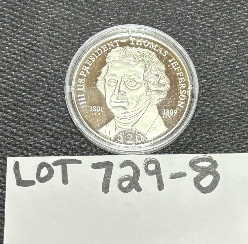 May include: A silver coin depicting Thomas Jefferson, with the inscription "III US PRESIDENT THOMAS JEFFERSON". The coin includes the years 1801 and 1809, and the value $20. Below the coin is a white paper with the text "LOT 729-8".