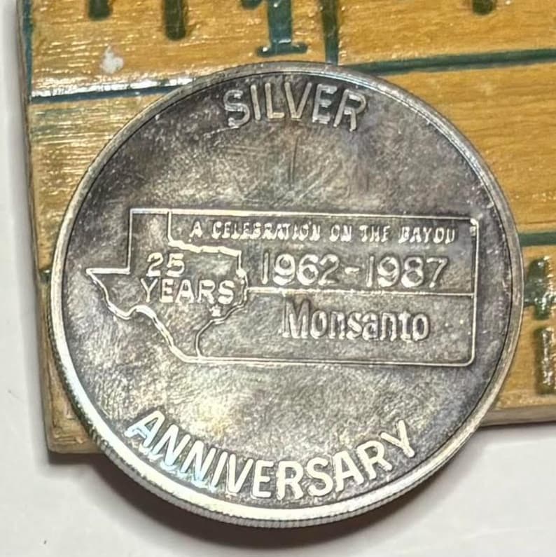 May include: Silver commemorative coin celebrating 25 years of Monsanto, 1962-1987. The coin features a map of the state of Louisiana with the text "A Celebration on the Bayou" and "Monsanto" inscribed.