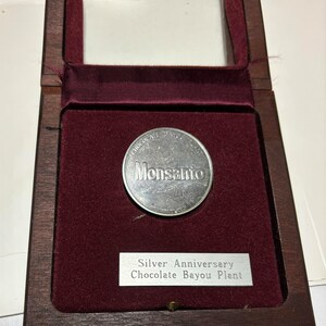 May include: A silver coin with the words "Chocolate Bayou Plant" and "Monsanto" on it, inside a wooden box with a red velvet lining. The box has a silver plaque that reads "Silver Anniversary Chocolate Bayou Plant".