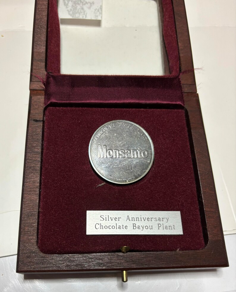 May include: A silver coin with the words "Chocolate Bayou Plant" and "Monsanto" on it, inside a wooden box with a red velvet lining. The box has a silver plaque that reads "Silver Anniversary Chocolate Bayou Plant".