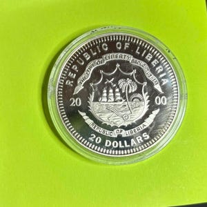 May include: A silver coin from the Republic of Liberia, in a clear plastic case, set against a vivid green backdrop. The coin's design includes a ship, palm trees, and the inscriptions "REPUBLIC OF LIBERIA" and "20 DOLLARS".
