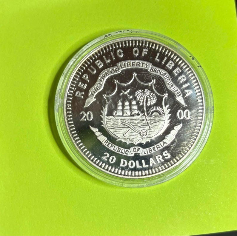 May include: A silver coin from the Republic of Liberia, in a clear plastic case, set against a vivid green backdrop. The coin's design includes a ship, palm trees, and the inscriptions "REPUBLIC OF LIBERIA" and "20 DOLLARS".
