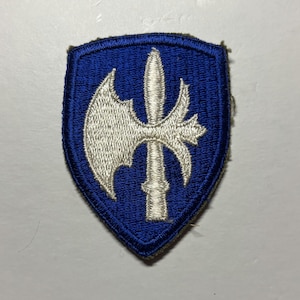 May include: A shield-shaped embroidered patch featuring a vibrant blue background with a white stitched design of a poleaxe. The patch has a thick, raised blue border that outlines the shield shape, with the white axe centered prominently.