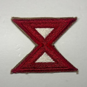 May include: An embroidered patch in the shape of an hourglass. The design features a vibrant red border and central cross-like structure, with two white triangular sections positioned at the top and bottom, all finished with a tan-colored edge.