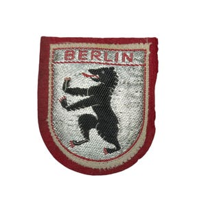 May include: A shield-shaped embroidered patch featuring a red border and a silver metallic background. The patch displays a black bear standing on its hind legs with red claws and tongue. The word BERLIN is stitched in red at the top.
