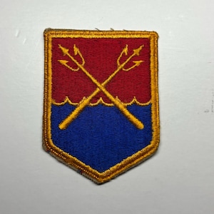 May include: A shield-shaped embroidered patch featuring a red upper section and a blue lower section separated by a wavy line. Two crossed yellow tridents are centered over the design, and the entire patch is bordered by a thick yellow thread.