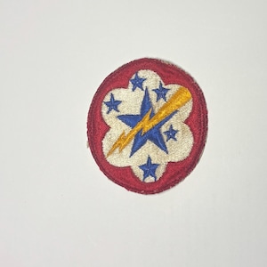May include: A circular embroidered patch featuring a red border surrounding a cream-colored center. The design includes a large blue star overlaid with a yellow lightning bolt, surrounded by five smaller blue stars arranged in a floral pattern.