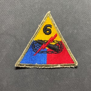 May include: A triangular embroidered patch featuring a yellow top section with a black number 6, a blue bottom left section, and a red bottom right section. A black tank tread graphic is centered with a red lightning bolt overlaying the design.