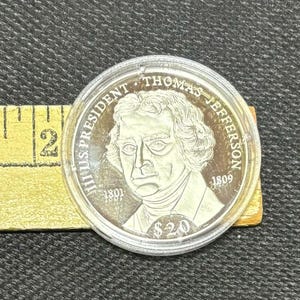May include: A silver coin depicting Thomas Jefferson, with the text "THOMAS JEFFERSON" and "$20". The coin is in a clear plastic case, with a yellow measuring tape visible in the background.