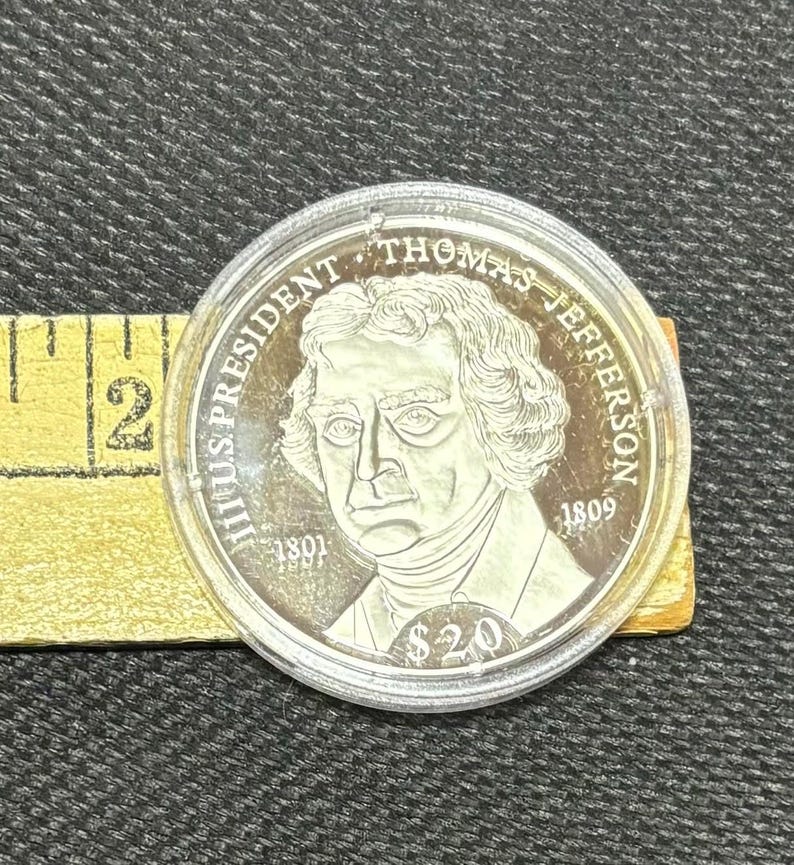 May include: A silver coin depicting Thomas Jefferson, with the text "THOMAS JEFFERSON" and "$20". The coin is in a clear plastic case, with a yellow measuring tape visible in the background.