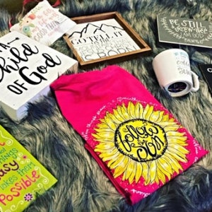 May include: An assortment of inspirational home decor items, including wooden signs with phrases like "I Am a Child of God" and "Give it to God and Go to Sleep." A pink towel with a sunflower design and a coffee mug are also visible.