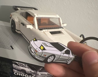 May include: A silver and black car-shaped enamel pin with yellow headlights. The pin is held in front of a white Lego car with a black spoiler. The text "CHOOSE YOUR" is visible on the surface behind the pin.