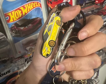 May include: A yellow Hot Wheels car keychain with black accents and a silver keyring. The keychain is held with other keys and a car key fob. In the background are several Hot Wheels car packages.