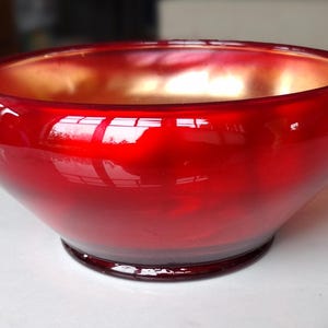 Puede incluir: Un cuenco de vidrio rojo brillante con interior dorado. La forma redondeada y la superficie lisa crean un contraste llamativo. Un objeto decorativo para el hogar.