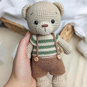 May include: A beige crocheted teddy bear toy featuring a striped green and white shirt and brown crocheted shorts with suspenders. The bear has two small round wooden buttons attached to the front of the suspenders and a dark brown stitched nose.