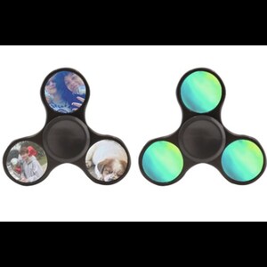 May include: Two black fidget spinners with different designs. One spinner has a photo of a dog and two people on the outer edges, and the other spinner has a green and blue gradient design.
