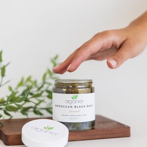 May include: A glass jar of Moroccan Black Soap with a white label. The label reads "arganier Moroccan Black Soap Unscented Organic Vegan Cruelty Free 4oz / 112 ml". The jar sits on a wooden tray with the lid beside it.
